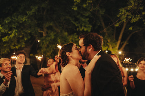 Bride and groom kiss under sparklers during outdoor evening wedding celebration