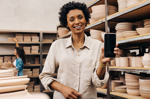 Smiling female ceramist showing a blank smartphone screen in her shop