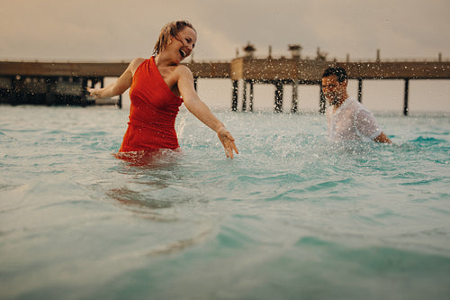 Retired couple splashing in the ocean, enjoying luxury in formal gowns near a pier at sunset