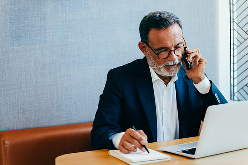 Senior businessman on phone and making notes at desk with laptop