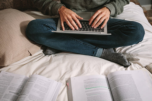 Man using laptop sitting on bed with books in front