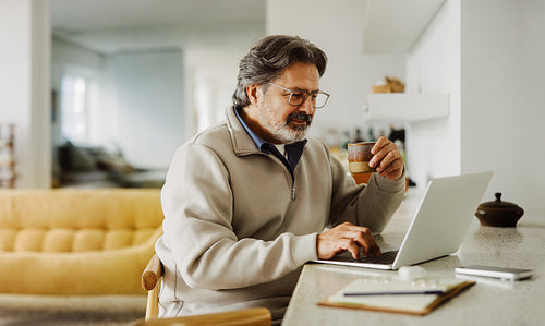 Senior man drinks coffee while browsing on a laptop