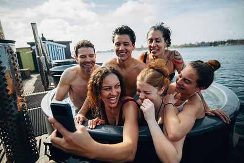 A social media moment features friends taking a selfie with a phone in an outdoor hot tub.