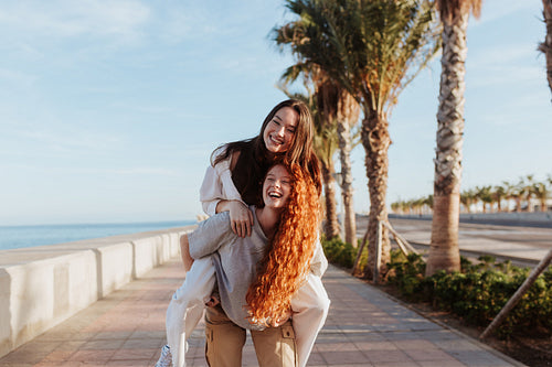 Playful young woman piggybacking her best friend in the promenad