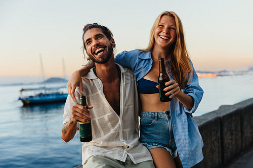 Two friends smiling by the water enjoying beers at sunset