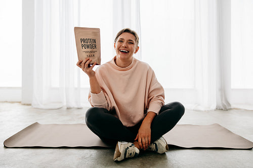 Woman holding protein powder while smiling during recovery on yoga mat