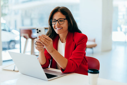Confident professional woman using a smartphone while working on a laptop