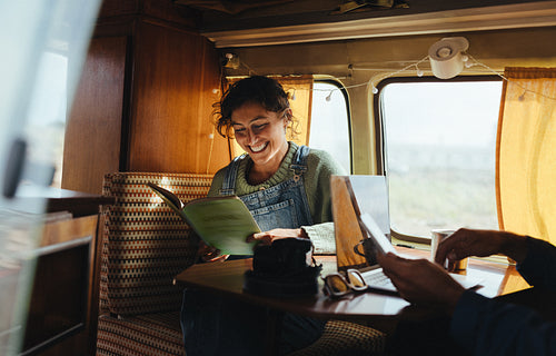 Inside a cozy camper, a woman reads a book while a friend works on a laptop