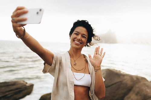 Smiling woman waving and taking a selfie by the seaside during vacation