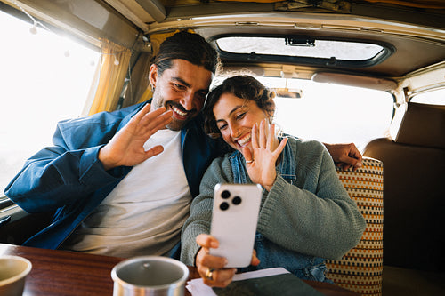 Happy couple on a road trip waving at smartphone on a video call inside a van
