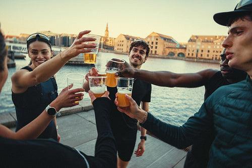Friends in a running group toast beer after a run by the waterfront at sunset