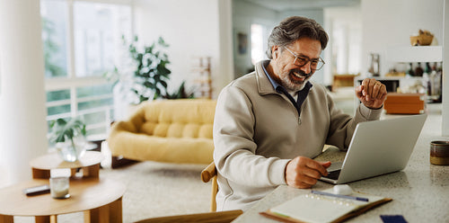Senior man uses laptop at home with wireless earbuds