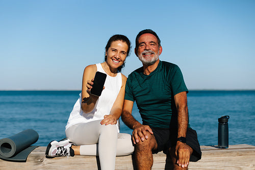 Senior couple outdoors showing a smartphone by a seaside backdrop