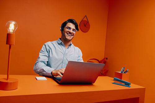 Male support agent with eyeglasses and headset working with laptop in bright office