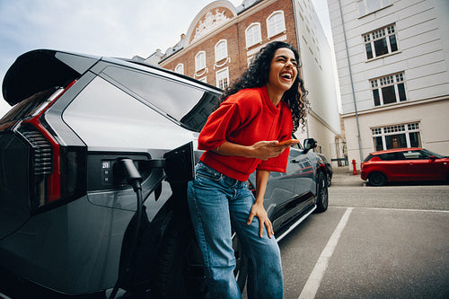 Woman charging electric car with smartphone at public station in urban setting
