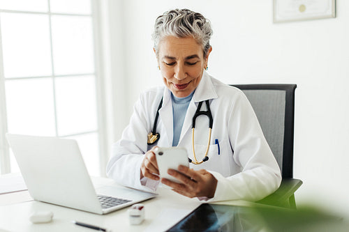 Mature doctor in a white lab coat using a smartphone while sitting in her office