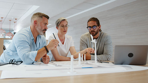 Working on a wind turbine design, three engineers discuss 3D models and drawings in an office
