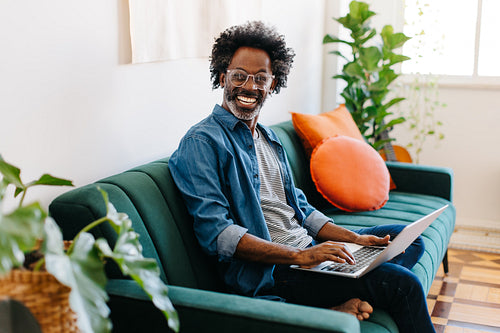 Afro blogger relaxing on couch, typing on a laptop in his living room