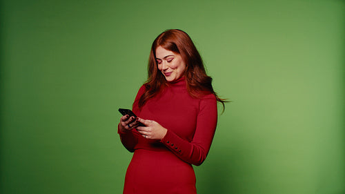 Smiling young woman with long red hair using phone against a green screen