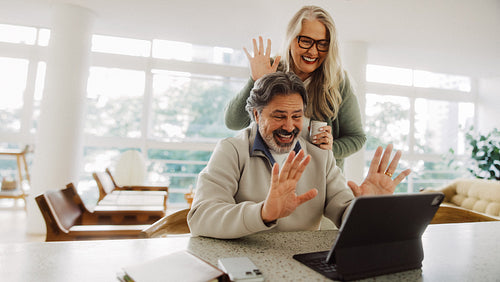 Laughing senior couple wave on a tablet in the kitchen