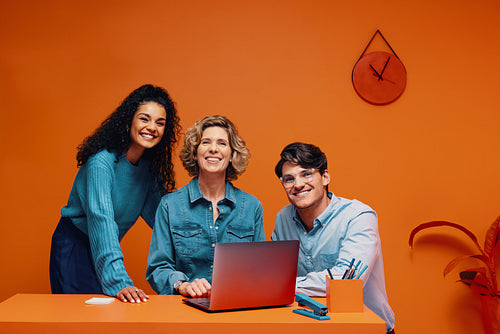 Diverse team of co-workers happily posing in vibrant office with laptop and colourful backgrounds