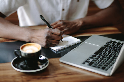Man writing notes sitting at a coffee shop with a laptop on the table