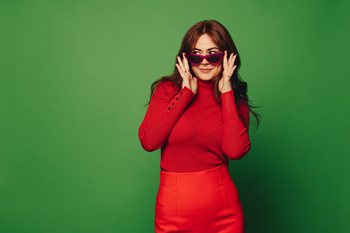 Vibrant fashionable woman standing on green background, smiling and peeking through sunglasses