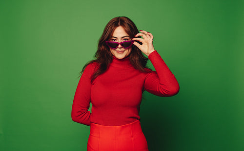 Vibrant woman peeking through sunglasses with stylish sweater on green background