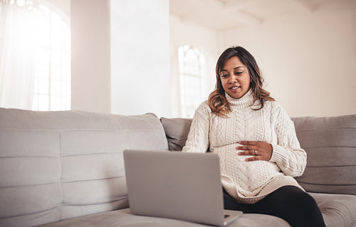 Beautiful pregnant woman with laptop sitting on sofa