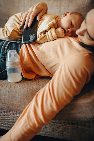 Mother rests with baby on couch, phone nearby.