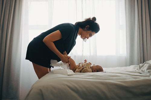 Mother changing baby’s diaper on bed in room