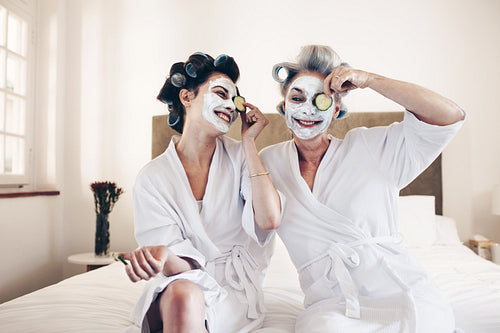 Happy women sitting on bed in bathrobes putting on beauty face mask