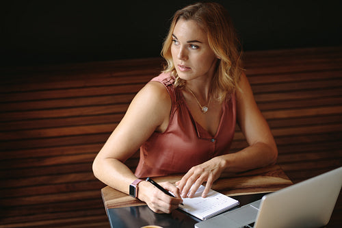 Woman writing notes sitting at a coffee shop with a laptop on the table