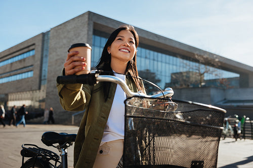 Woman with coffee walking with her bicycle through city plaza