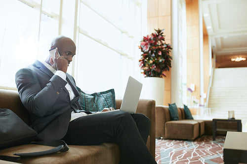 Businessman sitting in hotel lobby using cell phone and laptop