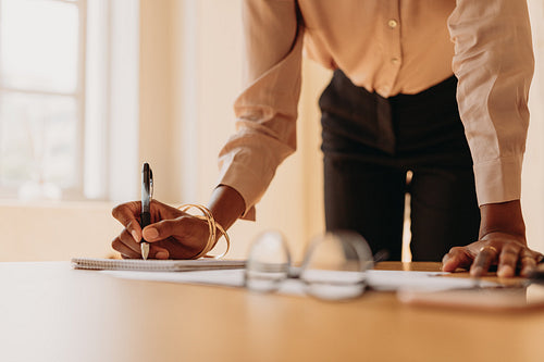 Businesswoman writing notes standing beside a table with hand resting on the table.