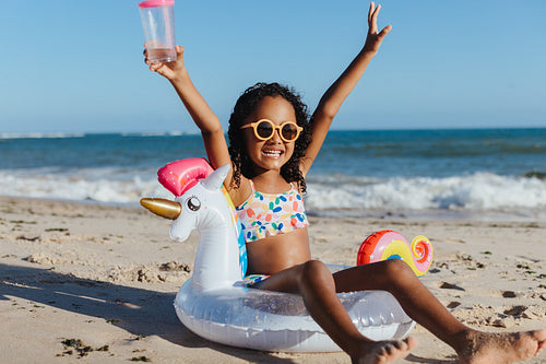Happy girl on holiday at the beach with inflatable unicorn floaty and drink