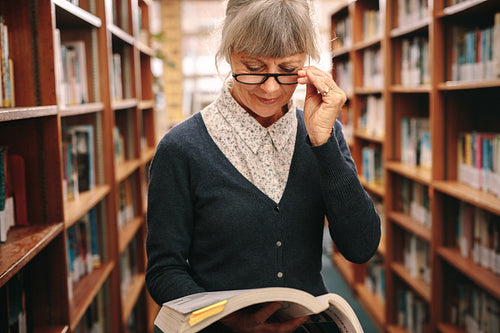 Portrait of a senior woman in eyeglasses reading a book in library