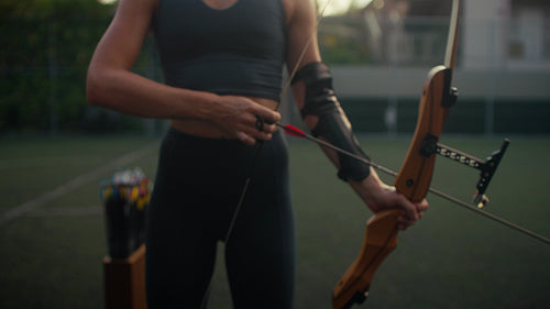 Young adult woman practicing archery, aiming with precision and focus outdoors at sunset