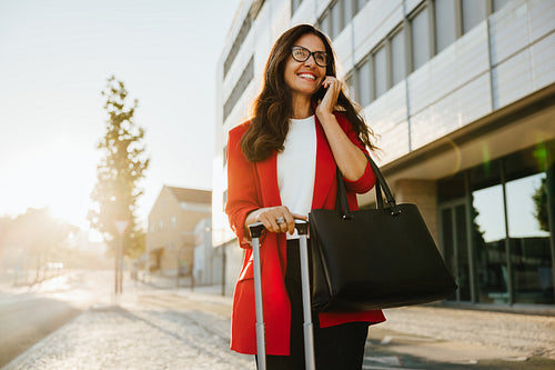 Smiling woman in red blazer carrying luggage and talking on phone outdoors