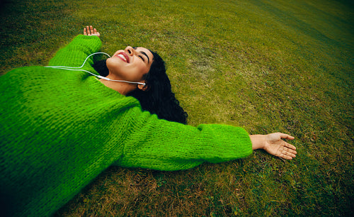 A woman with headphones listening to relaxing audio while laying on the grass outdoors