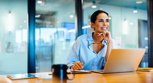 Smiling woman in office working on laptop and looking out with confidence