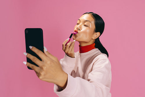 Selfie during makeup: Woman taking a picture while applying lipstick in a studio
