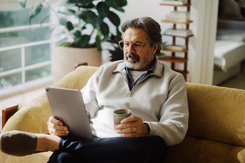 Reading senior man uses a tablet beside a mug at home