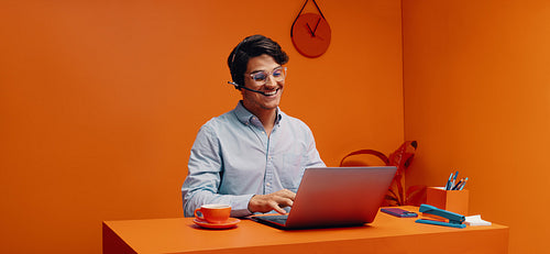 Entrepreneur wearing a headset while working on laptop in vibrant orange office