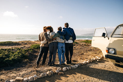 Friends stand together by the coast after a road trip, embracing near a van