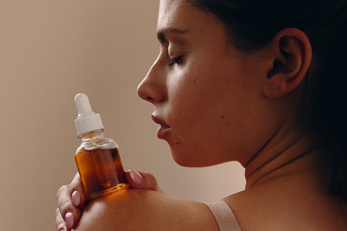 Woman holding facial oil bottle against shoulder, eyes closed