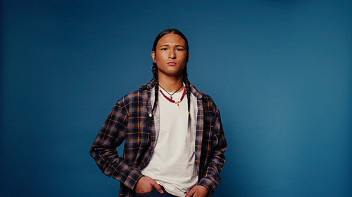Young man with braids and necklace poses seriously in a studio with blue background