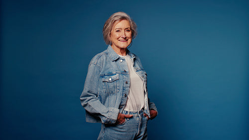 Confident senior woman smiles in denim jacket and jeans on blue background