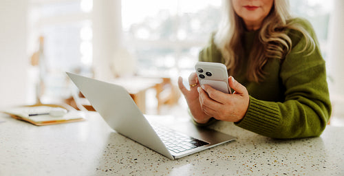 Senior woman uses smartphone beside laptop at home